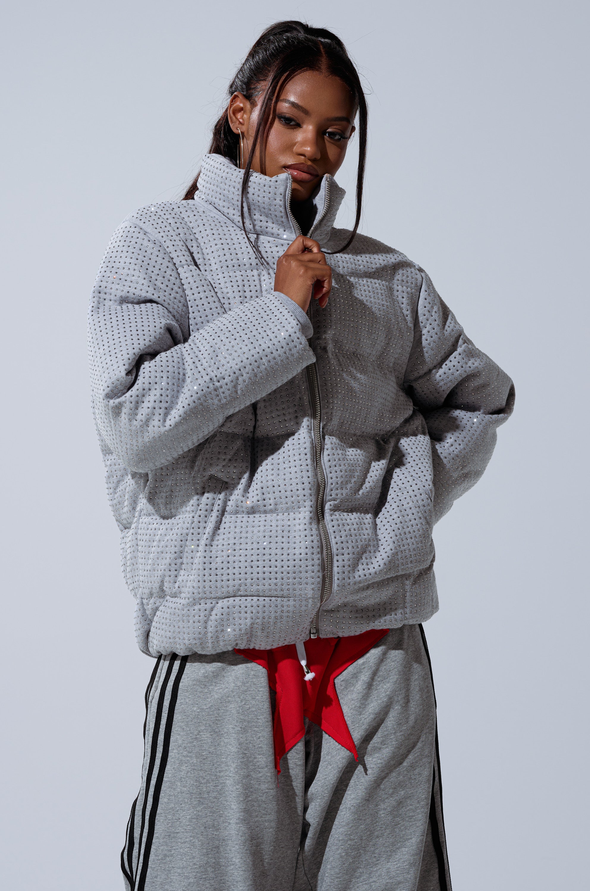A woman with long dark hair in pigtails poses against a plain light background, wearing the HOUDINI MID RHINESTONE PUFFER in light gray with a high collar, paired with gray striped sweatpants and a red top underneath.