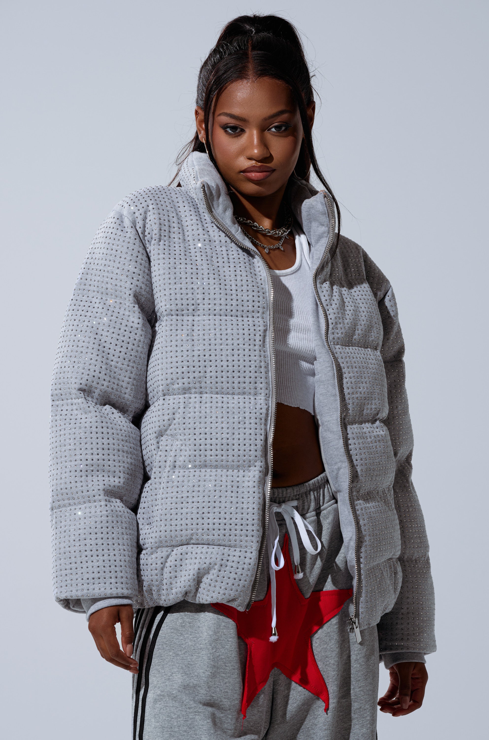 A woman wears the HOUDINI MID RHINESTONE PUFFER, a glittering silver-gray jacket, paired with a white crop top and gray sweatpants featuring bold red stars, standing confidently against a plain background.