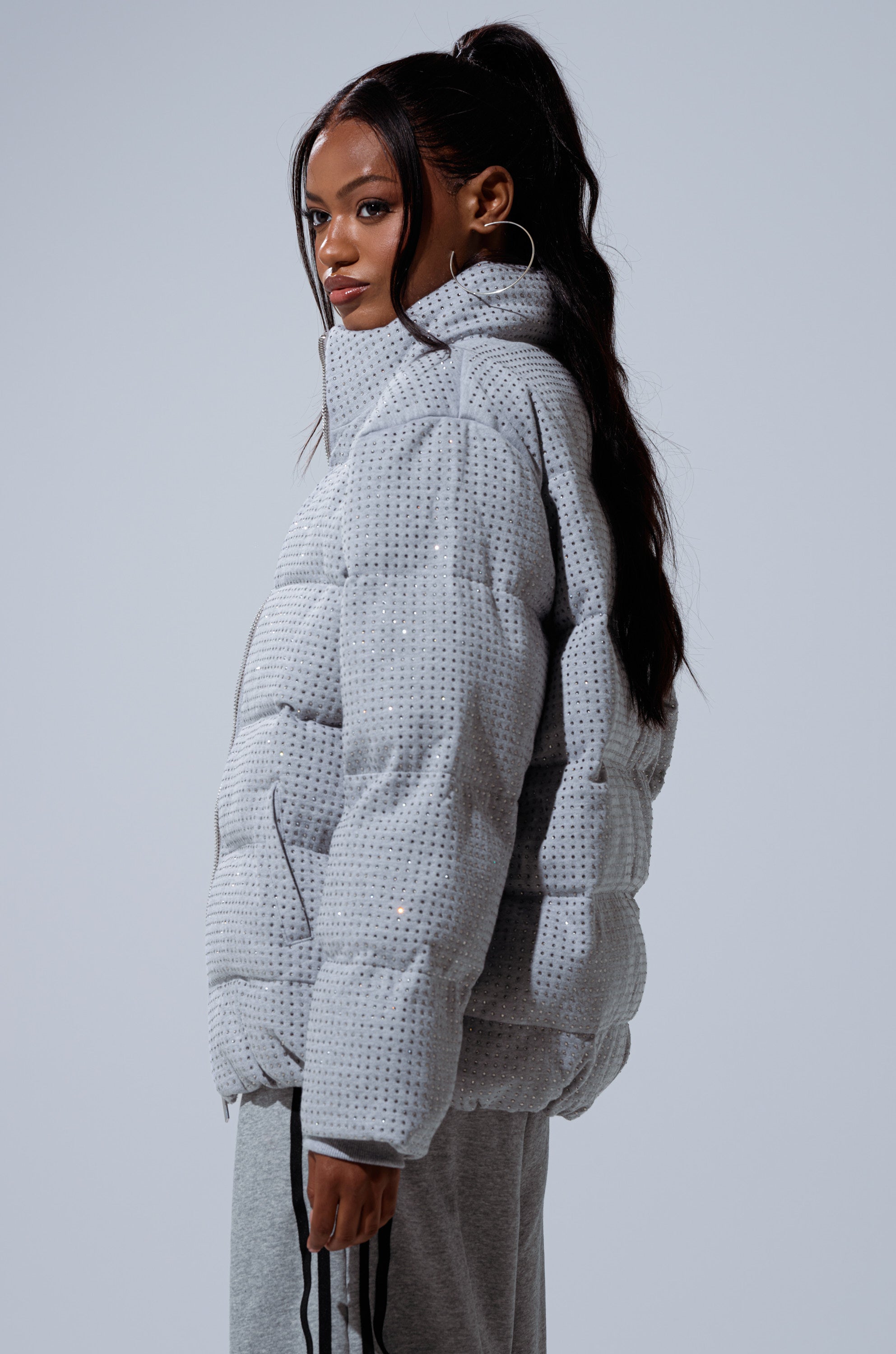 A woman with long dark hair in a high ponytail wears the HOUDINI MID RHINESTONE PUFFER and matching pants, standing sideways and looking at the camera against a plain light background.