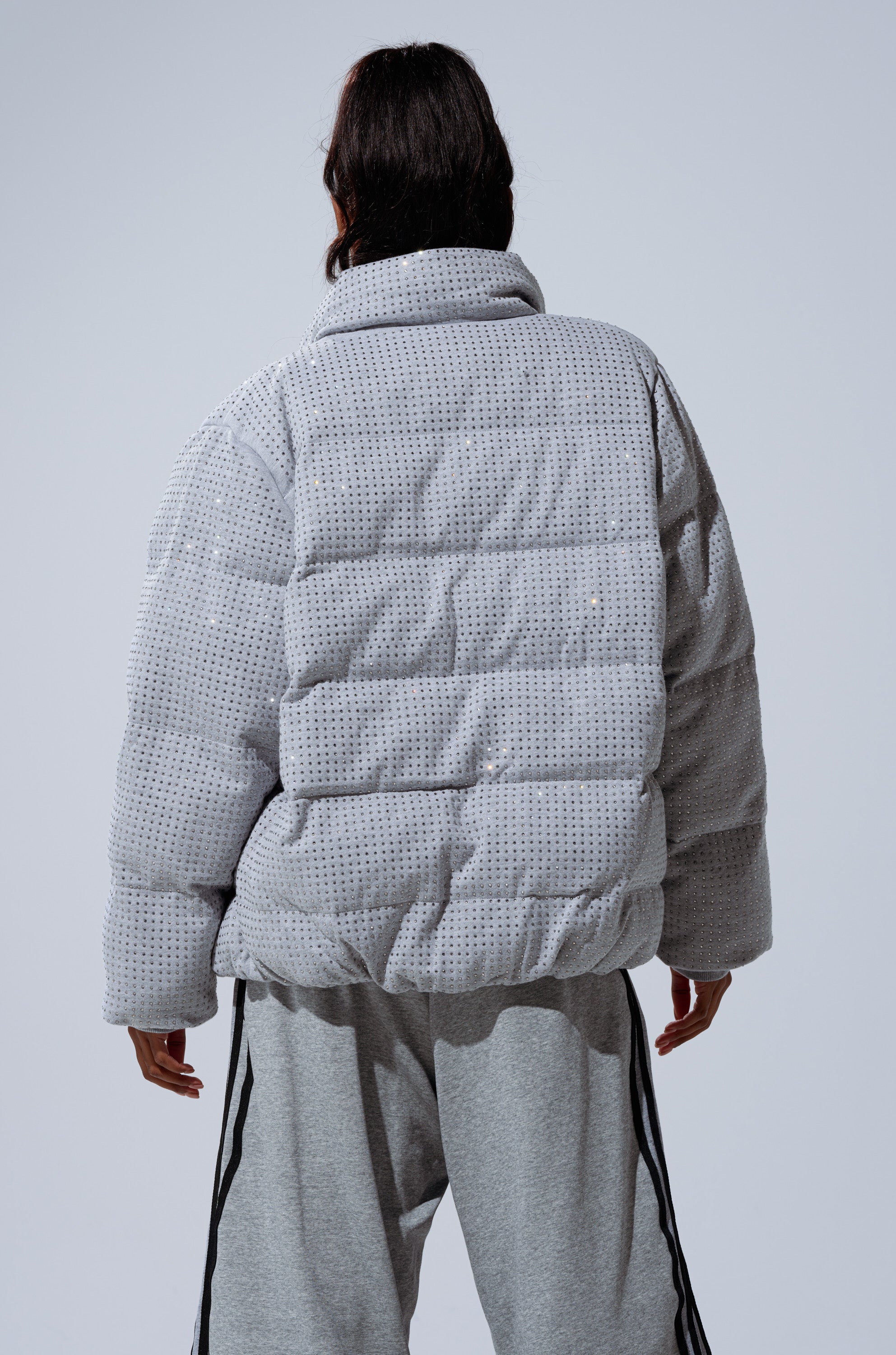 A person faces away from the camera wearing the HOUDINI MID RHINESTONE PUFFER, an oversized light gray jacket with a dotted pattern, paired with matching gray pants with black side stripes, against a plain background.