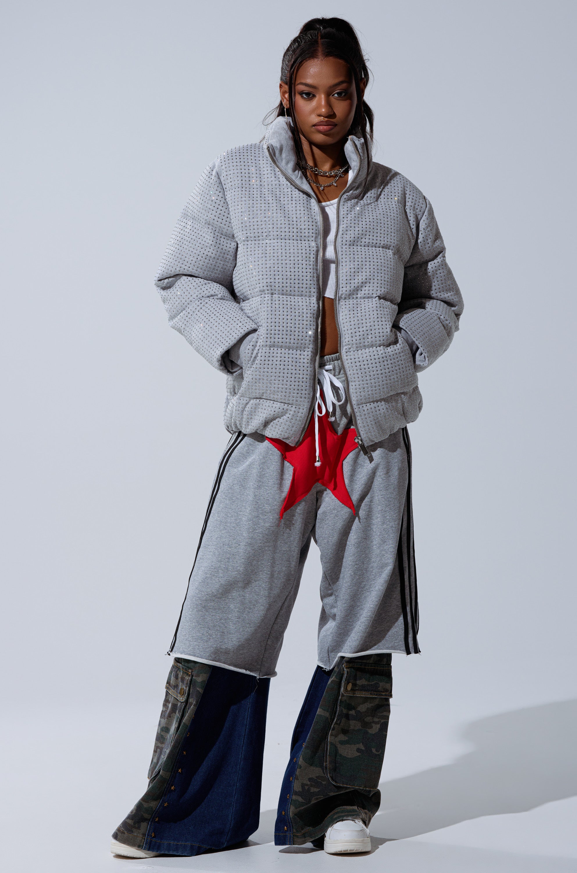 A woman stands confidently in the HOUDINI MID RHINESTONE PUFFER, gray athletic shorts with a red star accent, wide-leg denim and camouflage panel pants, white sneakers, and layered necklaces.