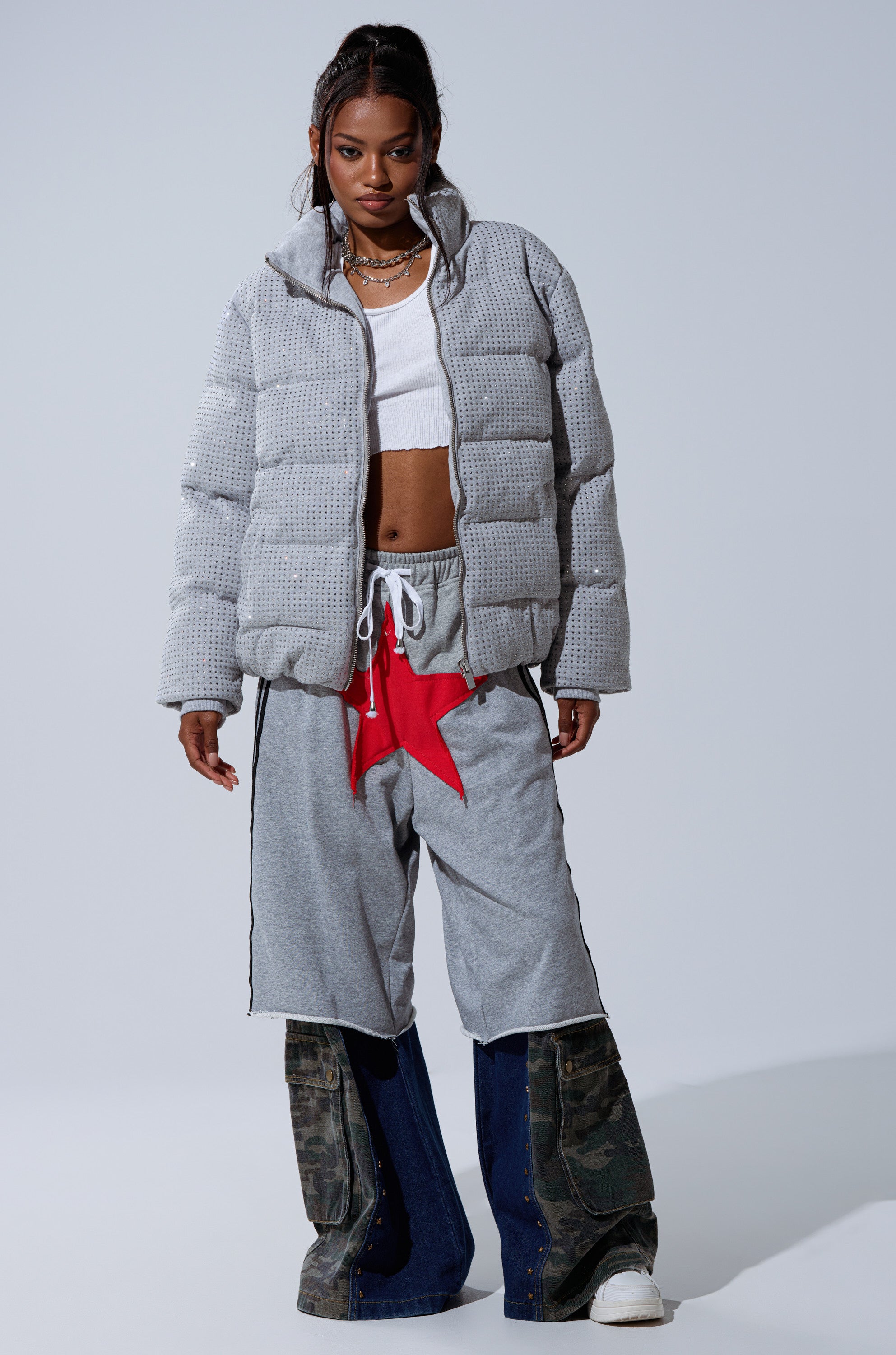 A woman poses in the HOUDINI MID RHINESTONE PUFFER, a cropped white top, gray drawstring star shorts, and layered camo panel pants over dark jeans. She accessorizes with necklaces and sports a high ponytail.