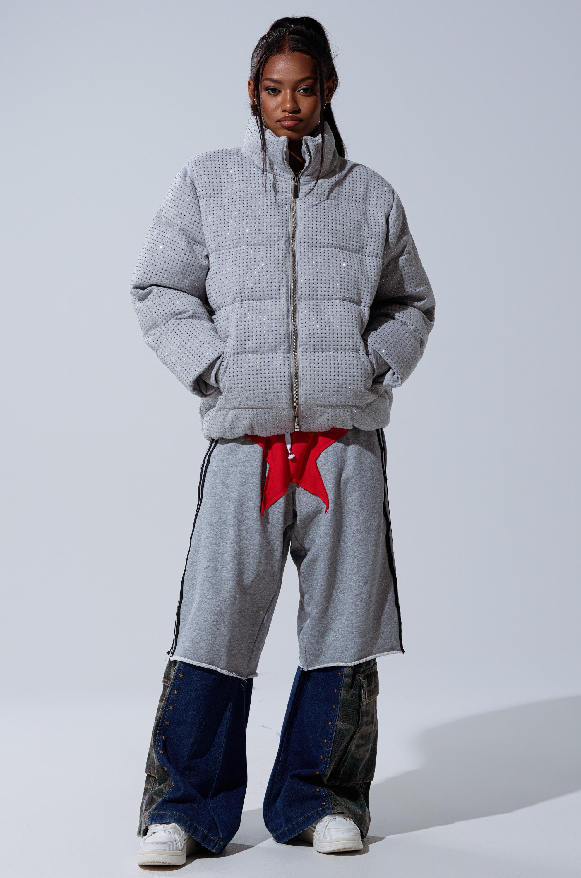 A person stands facing forward in the HOUDINI MID RHINESTONE PUFFER, light gray and oversized, paired with wide gray-blue layered pants, white sneakers, and a red accessory peeking out at the waist against a plain light background.