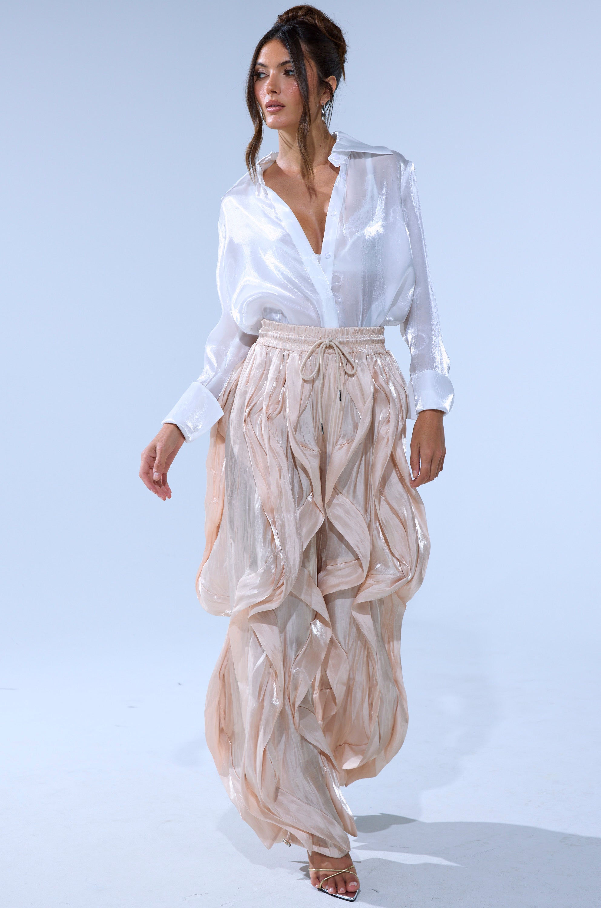 A woman confidently walks against a plain light background in the COOLEST ON THE BLOCK SATIN JOGGER, styled with an updo and strappy heels for a chic look.