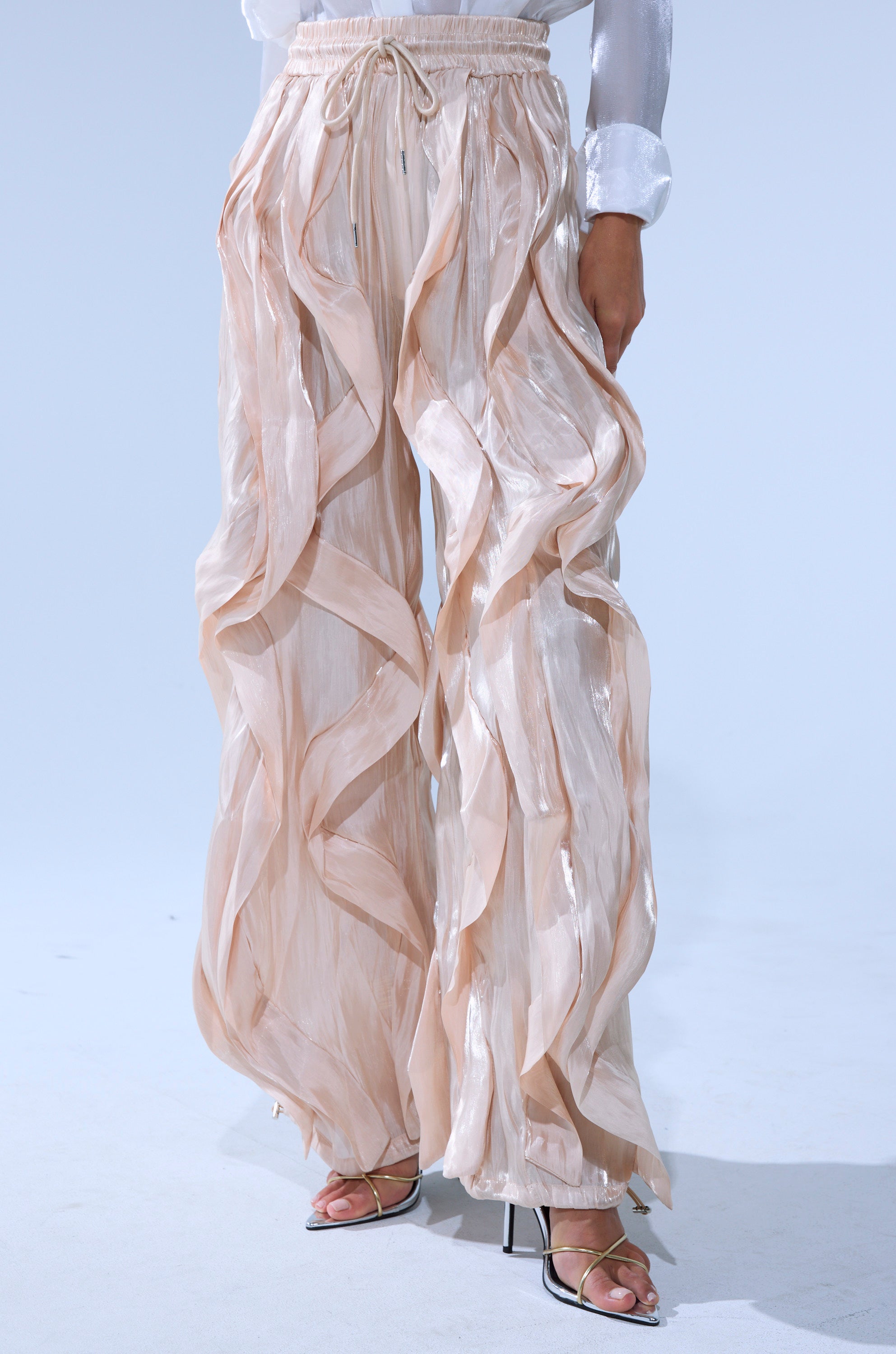 Wearing the COOLEST ON THE BLOCK SATIN JOGGER—wide-leg, sheer beige pants with ruffled, wavy details and an adjustable waistband—paired with strappy heeled sandals and a white long-sleeve blouse.