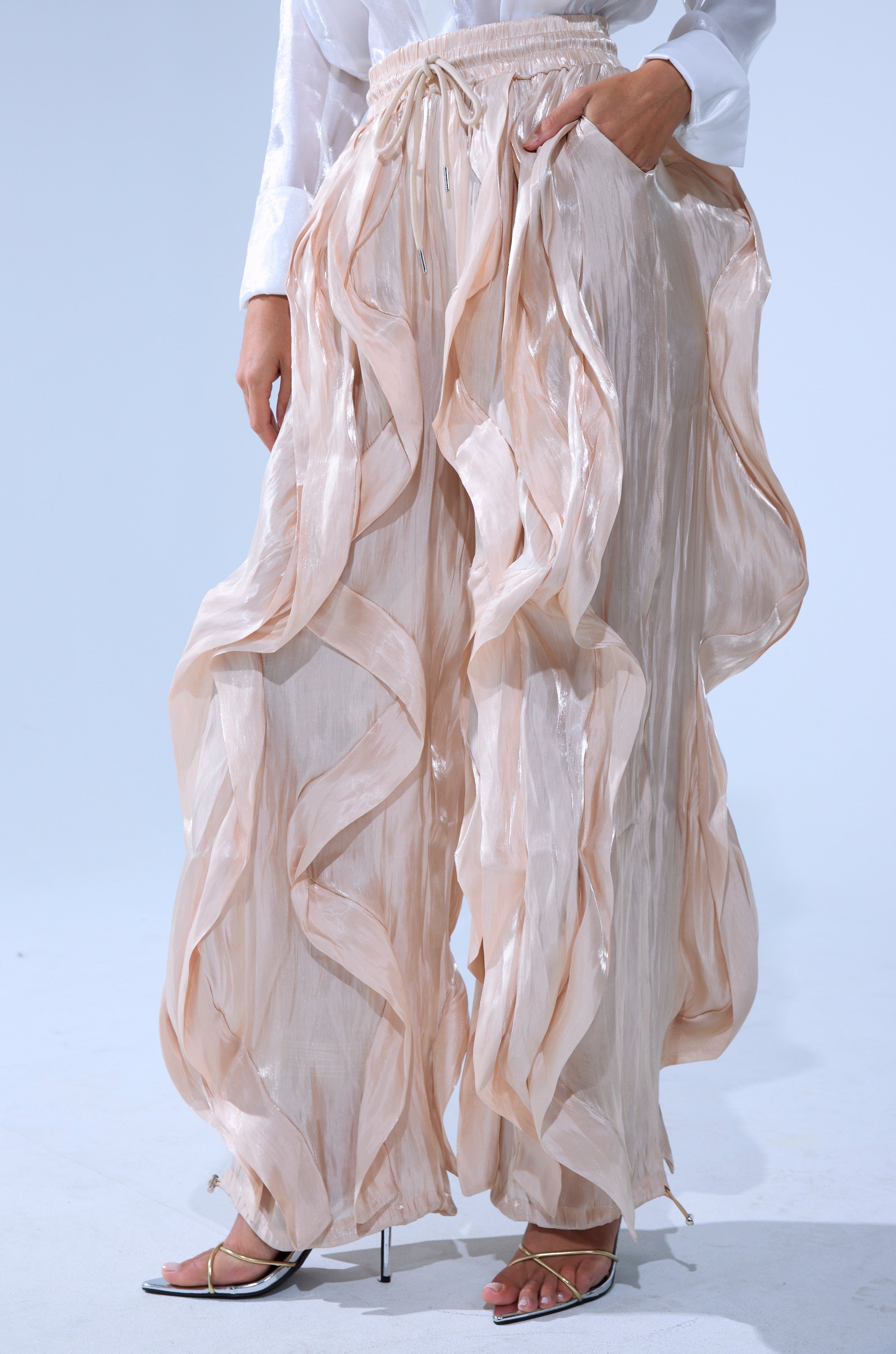 A person wearing the COOLEST ON THE BLOCK SATIN JOGGER—shiny, sheer, cream-colored ruffle pants with a wide-leg fit and elastic waistband—paired with white heeled sandals and a white long-sleeve top.