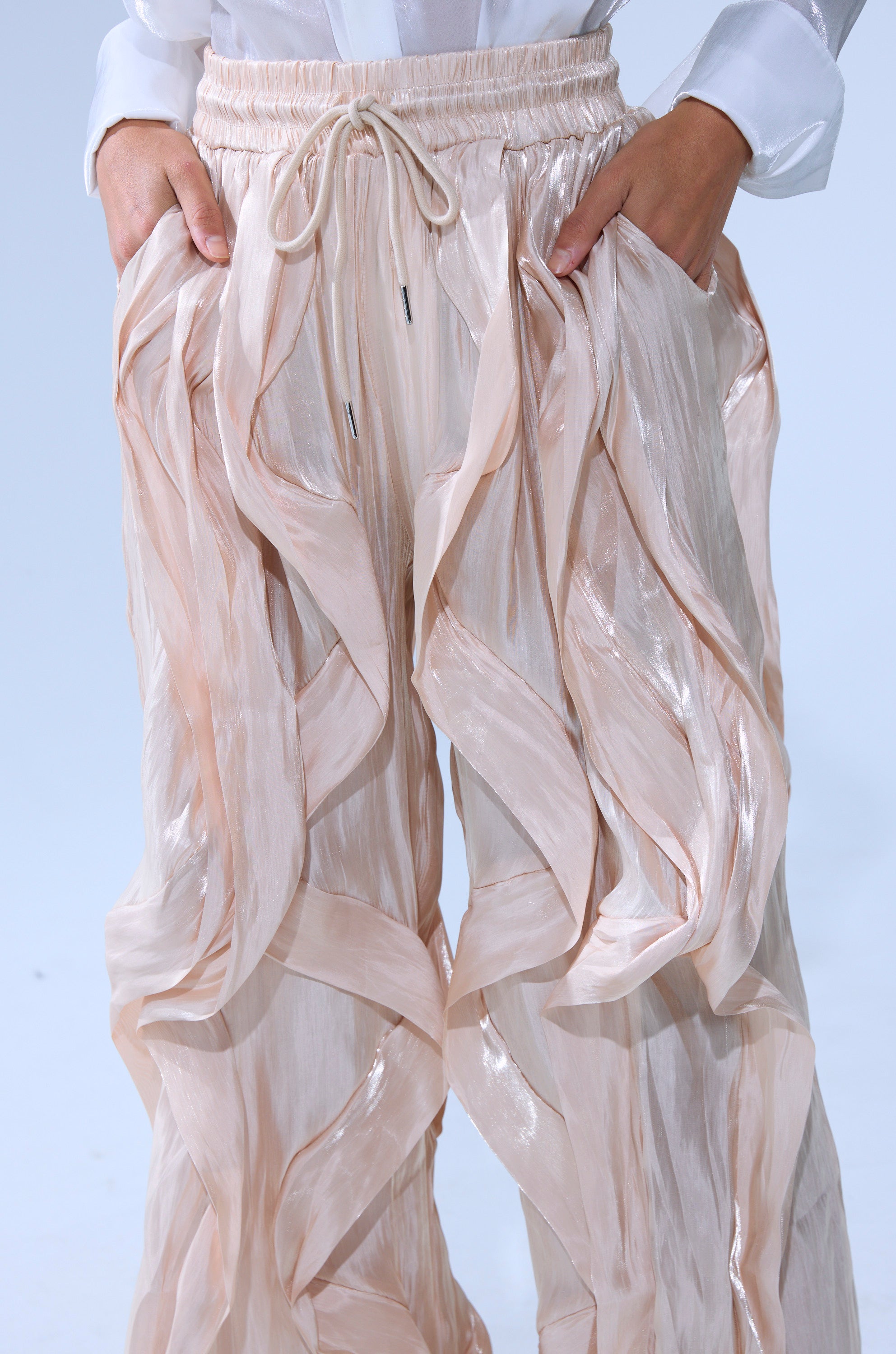 Wearing the COOLEST ON THE BLOCK SATIN JOGGER in shiny light beige with wavy ruffle details and an adjustable waistband, paired with a white long-sleeve top; the relaxed wide-leg fit suits their hands-in-pockets pose.