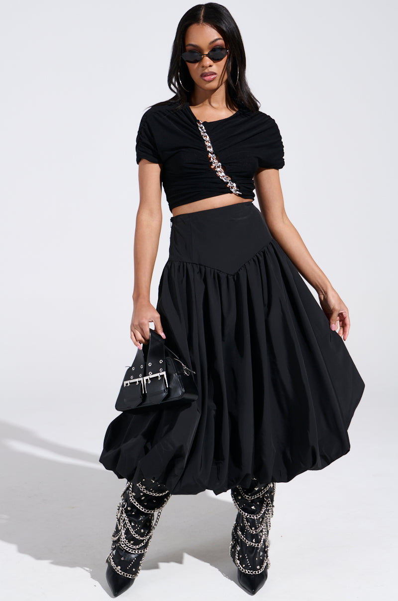 A woman in sunglasses wears a black ruched crop top and the chic I FOUND YOU DROP WAIST MIDI SKIRT IN BLACK. She completes her stylish look with a black handbag and chain-adorned black boots, set against a plain white background.