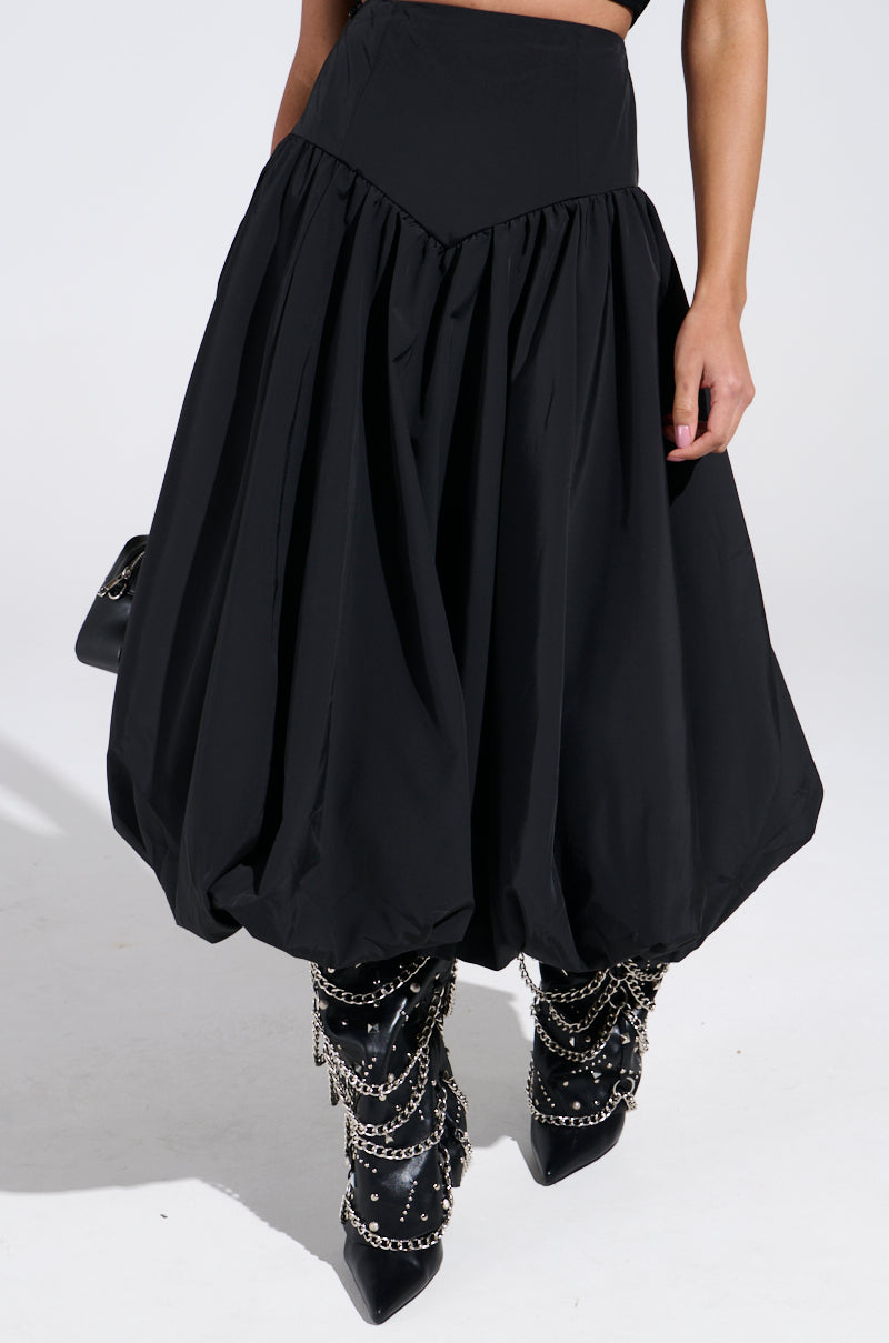 A person wears the I FOUND YOU DROP WAIST MIDI SKIRT IN BLACK paired with black pointed boots with silver chains, set against a plain white background. The bold skirt partially obscures their upper body beneath its voluminous fabric.