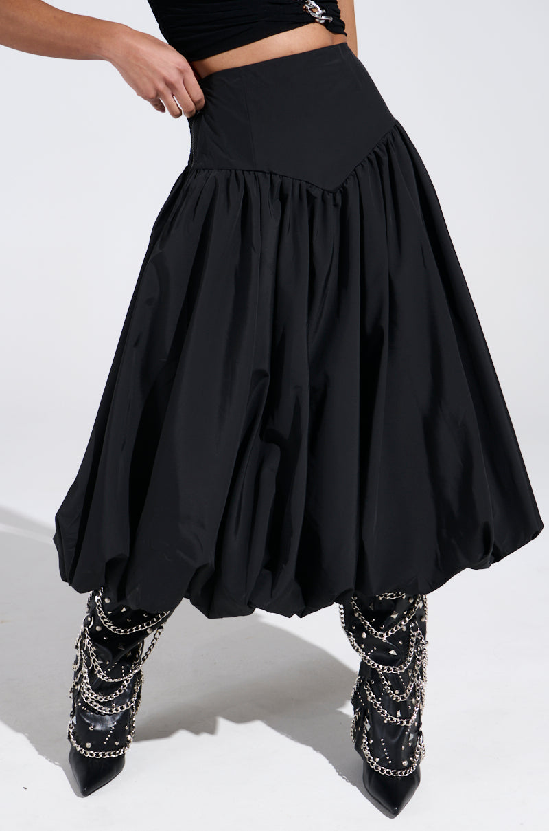 The image artistically shows the lower half of a person wearing an AKIRA Label's I FOUND YOU DROP WAIST MIDI SKIRT IN BLACK paired with pointed black boots featuring silver chain details, set against a plain background.