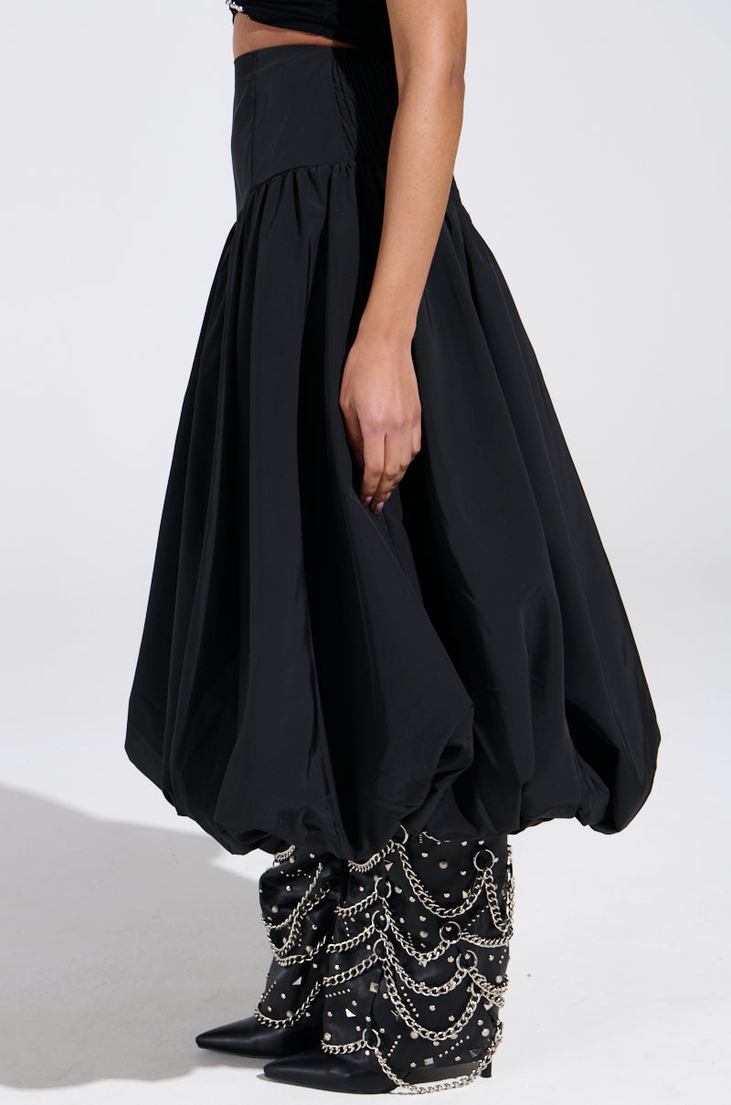 A person is wearing the "I FOUND YOU DROP WAIST MIDI SKIRT IN BLACK" from AKIRA, paired with pointy-toed boots accented with silver chains and studs. The ensemble stands out against a plain white background.