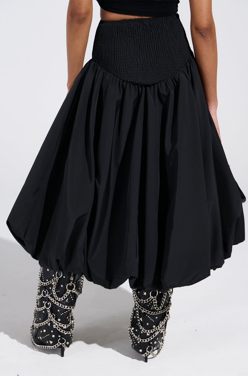 An individual poses with their back to the camera against a light gray backdrop, wearing the I FOUND YOU DROP WAIST MIDI SKIRT IN BLACK. The voluminous skirt flows gracefully, paired with ornate black boots adorned with chains.