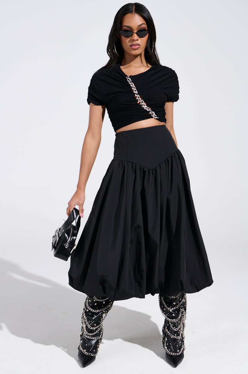 A woman in stylish attire poses confidently in a trendy I FOUND YOU DROP WAIST MIDI SKIRT IN BLACK from the AKIRA Label. She wears a ruched crop top, voluminous skirt, and chain-adorned boots, accessorized with oval sunglasses and a small black purse with a chain strap against a white background.