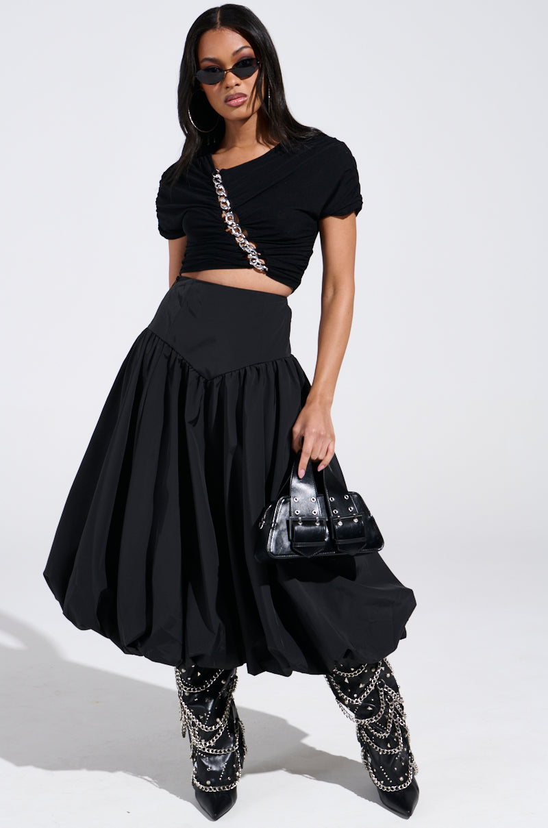 A woman in a stylish black outfit exudes confidence, wearing an "I FOUND YOU DROP WAIST MIDI SKIRT IN BLACK" paired with a cropped top and pointed, chain-adorned boots. Her ensemble is accessorized with a small handbag and sunglasses, the neutral background enhancing her bold fashion statement.