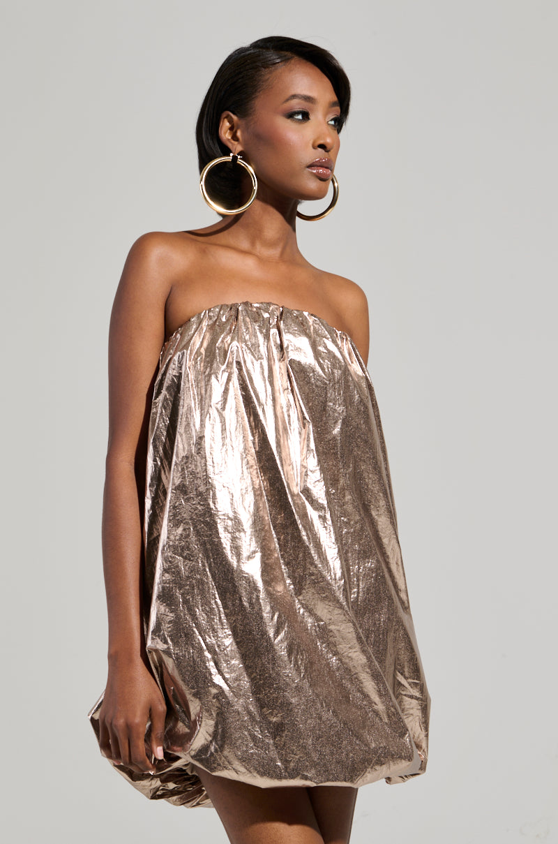 A woman stands against a plain background, wearing the STOPPING TRAFFIC GOLD MINI DRESS, featuring an irregular hem. She has large hoop earrings and her hair is styled short and sleek. She gazes into the distance with a confident expression.