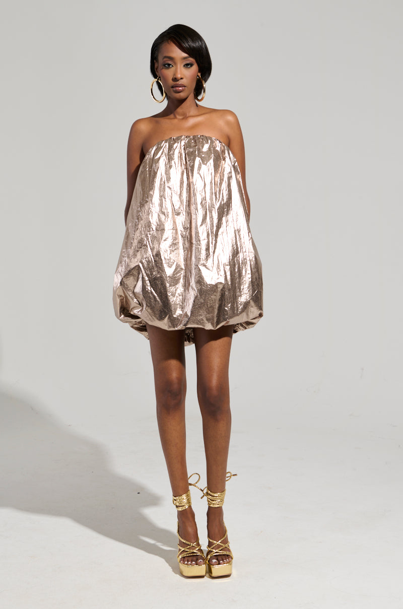 A woman stands confidently in the STOPPING TRAFFIC GOLD MINI DRESS, which features a metallic knit design that resembles a bubble shape. She pairs it with large hoop earrings and golden platform heels. The plain gray background enhances her bold style.
