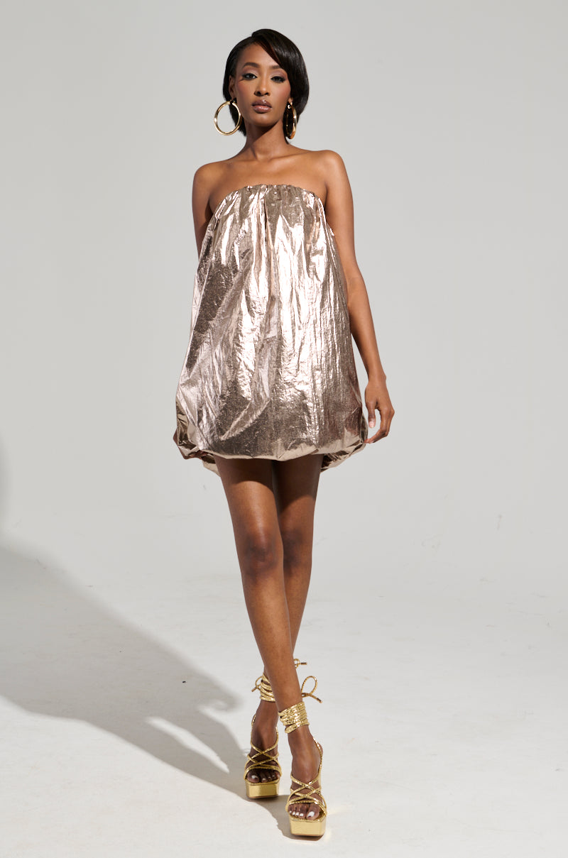 A woman poses in the STOPPING TRAFFIC GOLD MINI DRESS, showcasing its ruched metallic knit texture, strapless design, and bubble hem. She enhances her chic look with large hoop earrings and gold platform heels. The plain, well-lit background highlights her elegant outfit and poised stance.