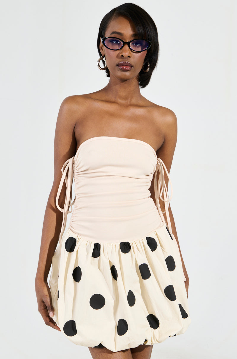 A person is flaunting the ON TREND BUBBLE MINI DRESS, which features a strapless, ruched beige top with tie details and a voluminous skirt in a stylish bubble hem design adorned with large black polka dots. They complete the outfit with sunglasses and chin-length hair.