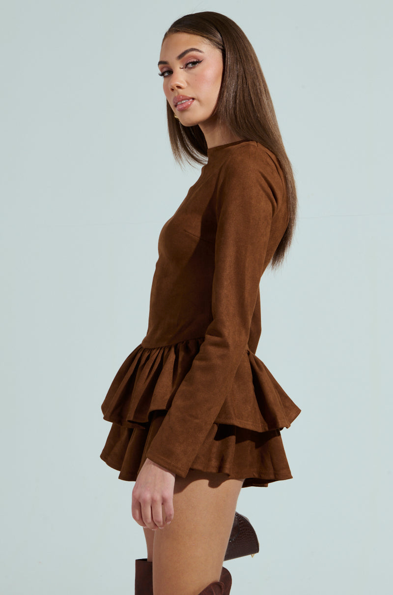 A woman with long brown hair stands in profile against a light background, wearing a brown SUEDE RUFFLE LONG SLEEVE ROMPER. She has a neutral expression and is looking at the camera.