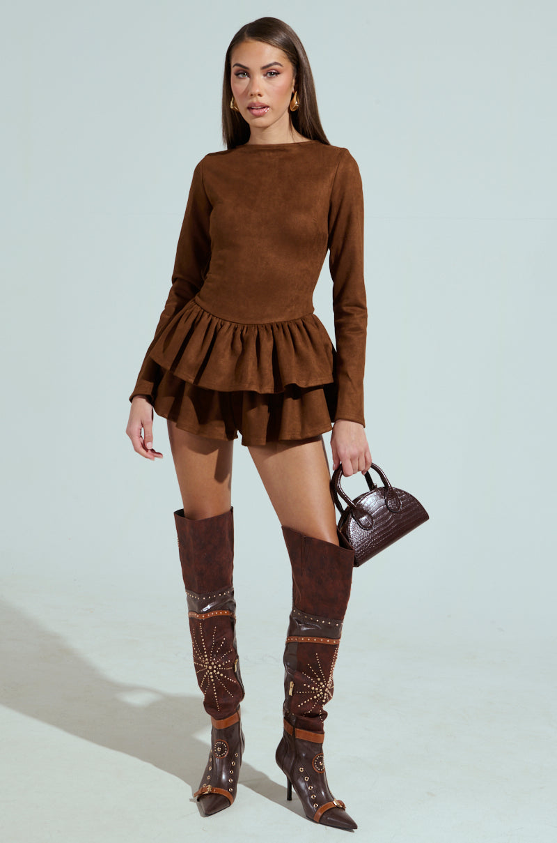 A woman dons the Suede Ruffle Long Sleeve Romper, featuring a scoop neckline and ruffled hem, paired with tall patterned brown boots. She holds a small brown handbag against a plain white background.