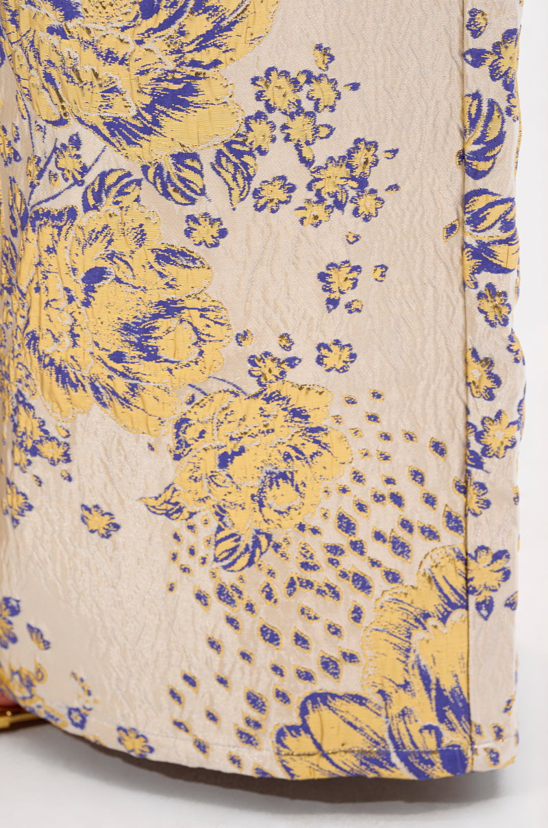 Close-up of GOLDIE BROCADE TROUSER fabric, showcasing a textured surface with a floral brocade pattern in yellow and blue on pale beige, featuring large flowers, small blossoms, and dotted accents.