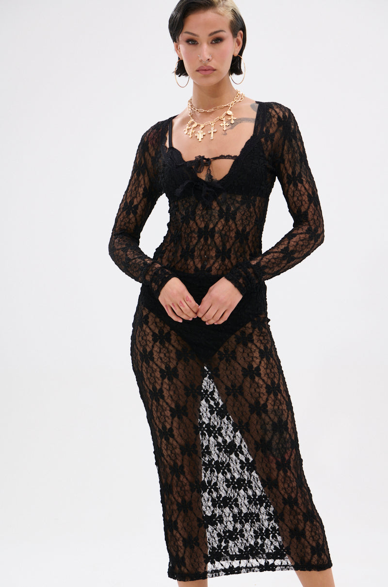 A woman models the LIZZIE LACE MAXI DRESS—a black, bodycon maxi with floral lace, deep neckline, and high slit—against a white background, accessorized with a chunky gold necklace and hoop earrings for an elegant touch.