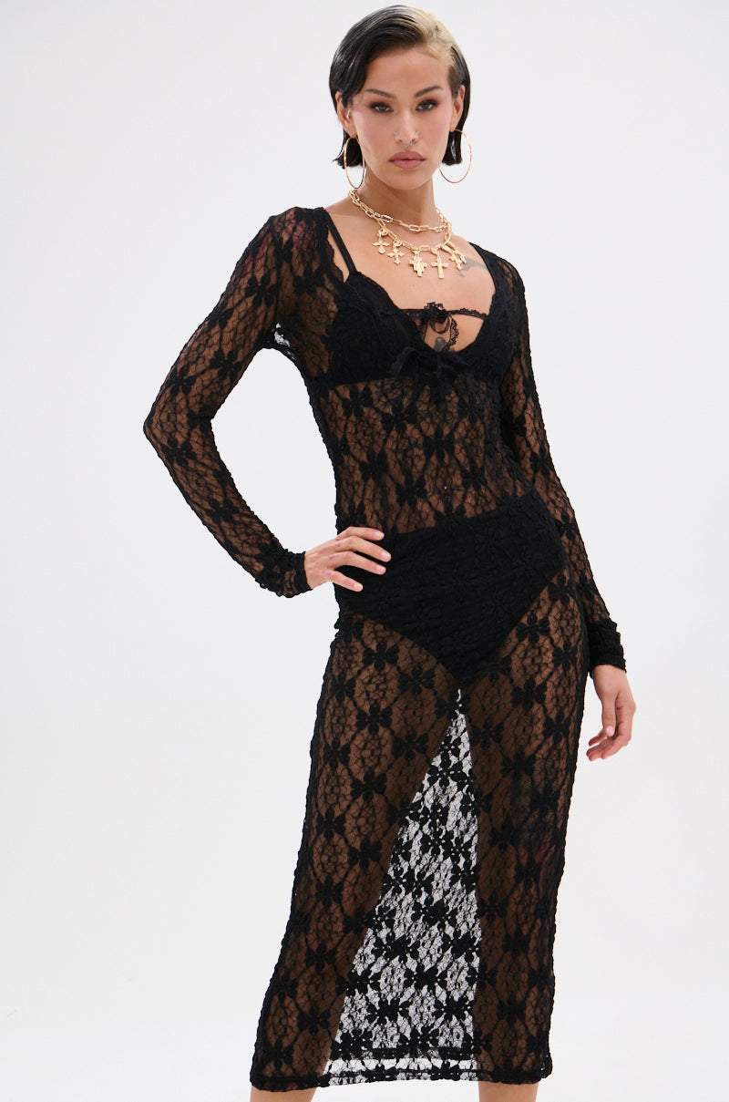 A woman poses confidently in the LIZZIE LACE MAXI DRESS—a black sheer mesh, long-sleeved dress with a floral lace base over black undergarments—accessorized with large hoop earrings and a gold statement necklace, against a white background.