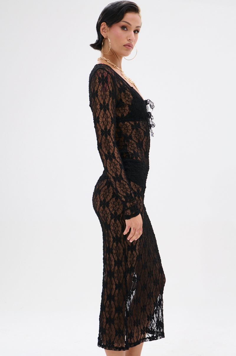 A woman with short dark hair and large hoop earrings stands in profile against a white background, wearing the LIZZIE LACE MAXI DRESS—a sheer mesh, long-sleeve maxi dress with a floral lace base over a black slip.