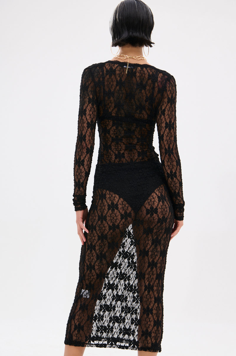 A person with short, dark hair faces away in the LIZZIE LACE MAXI DRESS, featuring sheer mesh, a floral lace base, back slit, and revealing black undergarments against a plain light background.