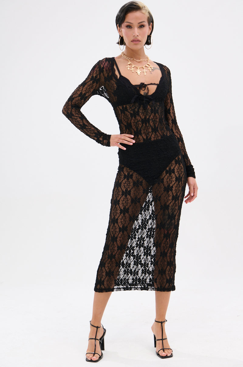 A woman models the LIZZIE LACE MAXI DRESS, a sheer mesh, long-sleeve, mid-calf dress featuring a scoop neckline and high side slit. She pairs it with black undergarments, strappy sandals, and a gold statement necklace.