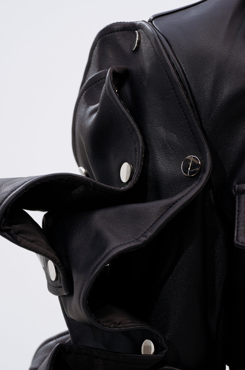 A close-up of the SNAP OUT OF IT ULTRA CROP MOTO jacket in black faux leather displays silver snap buttons, layered folds, sculptural sleeves, and detailed hardware against a simple light background.