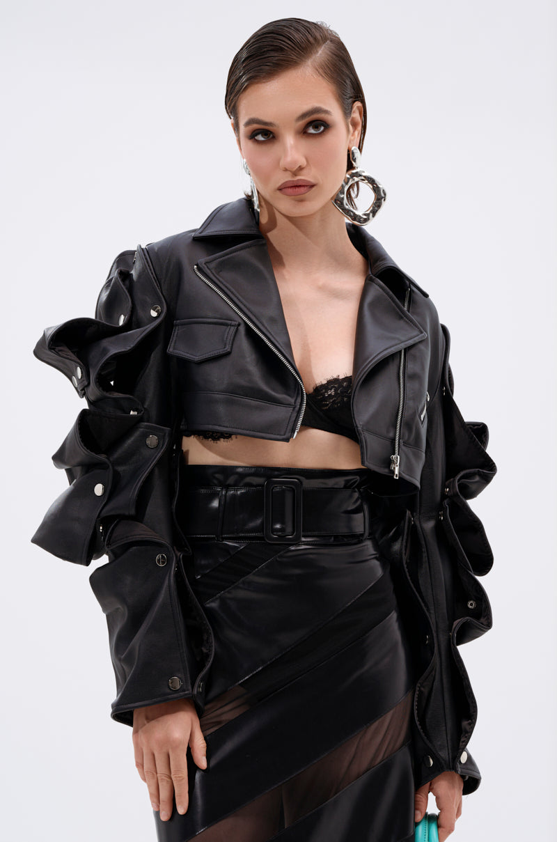 A woman with short, slicked-back hair wears the SNAP OUT OF IT ULTRA CROP MOTO jacket with sculptural sleeves over a black bralette and high-waisted black skirt with sheer panels, accessorized with large hoop earrings.