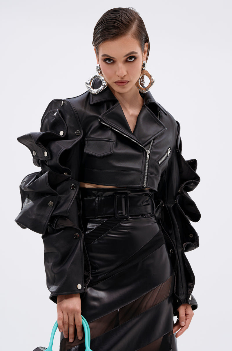 A woman stands confidently against a white background, wearing the SNAP OUT OF IT ULTRA CROP MOTO jacket with sculptural ruffled sleeves and a matching high-waisted skirt, holding a turquoise-handled bag.