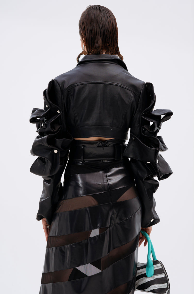 A person with wet, slicked-back hair wears the SNAP OUT OF IT ULTRA CROP MOTO jacket and a high-waisted black skirt with sheer panels, holding a turquoise and gray patterned bag against a plain white background.