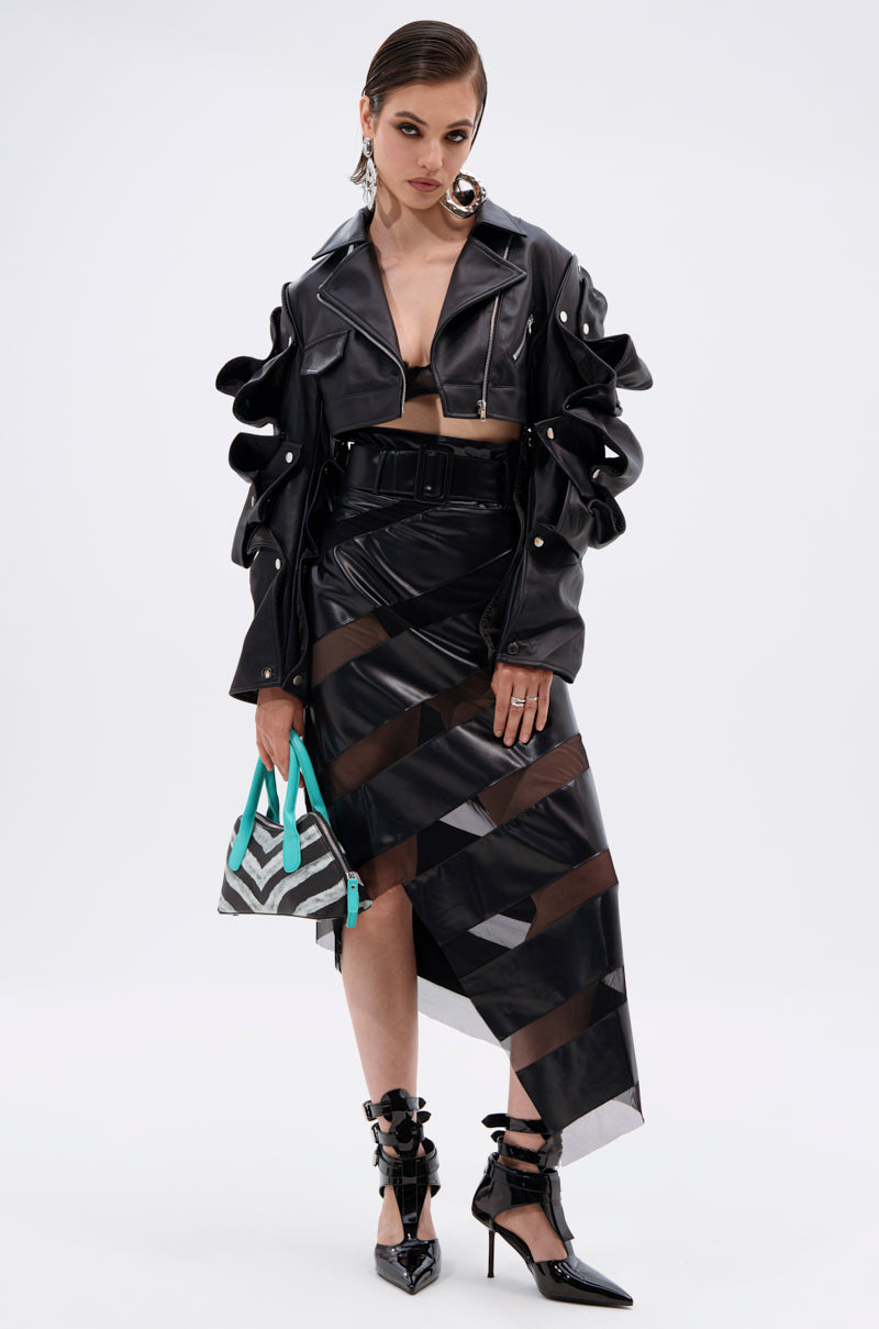 A woman wears the SNAP OUT OF IT ULTRA CROP MOTO jacket with ruffled sleeves, a black bra top, an asymmetrical black skirt with sheer striped panels, black heels, and holds a geometric turquoise handbag.
