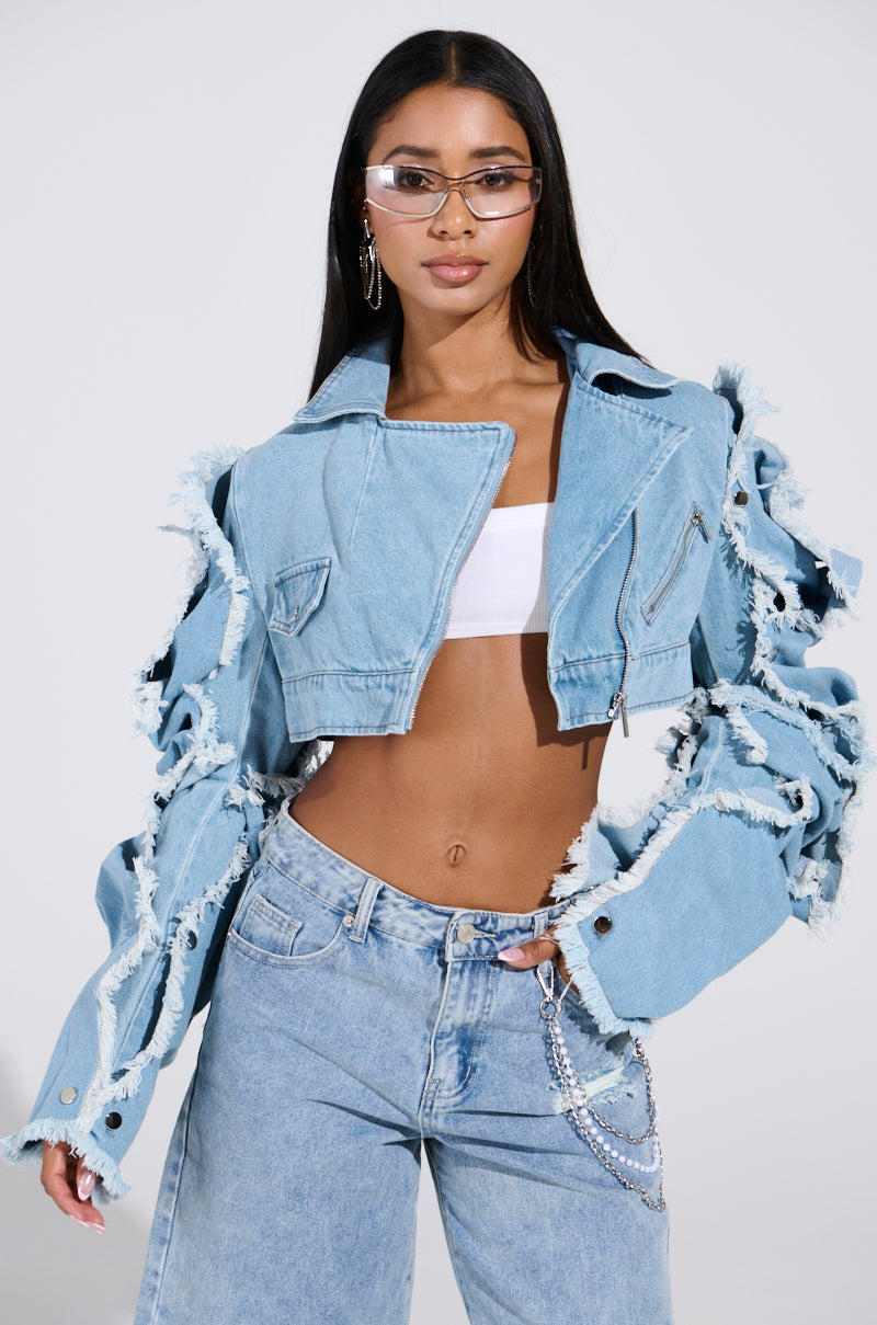 A person confidently poses in the SNAP OUT OF IT ULTRA CROP MOTO IN DENIM, featuring frayed edges and a light wash. They're styled with matching jeans, long hair, clear glasses, and a white cropped top against a plain light-colored background.