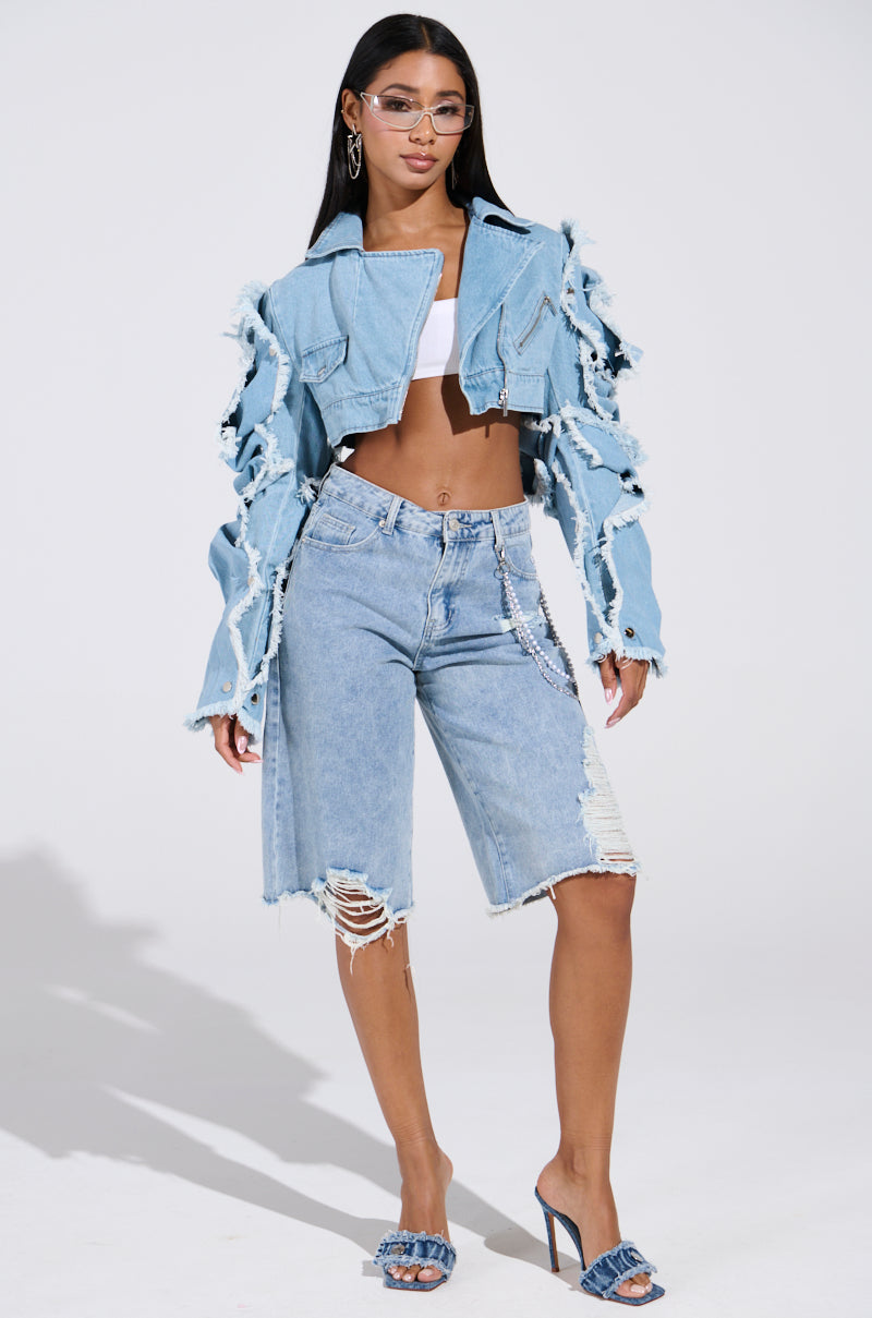 A woman poses confidently in a torn light wash SNAP OUT OF IT ULTRA CROP MOTO denim jacket and long denim shorts, revealing a white crop top beneath. She accessorizes with glasses, hoop earrings, and heeled sandals against a plain background.