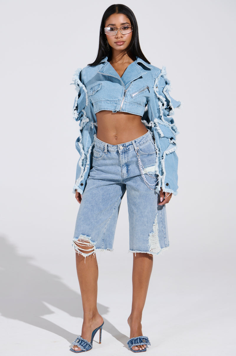 Showcasing confidence in the "SNAP OUT OF IT ULTRA CROP MOTO IN DENIM," a person stands in distressed denim jacket and blue knee-length shorts. With long hair, sunglasses, and open-toed heels, they exude effortless style against a plain backdrop.