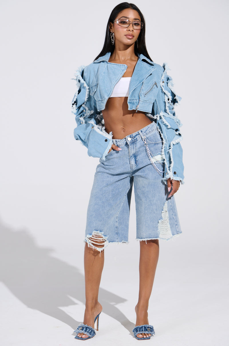 A person poses confidently in an ultra-cropped fit, wearing a distressed denim jacket and knee-length denim shorts. Paired underneath is the chic SNAP OUT OF IT ULTRA CROP MOTO IN DENIM. Silver chain and sunglasses complement, while light wash denim sandals complete the ensemble.