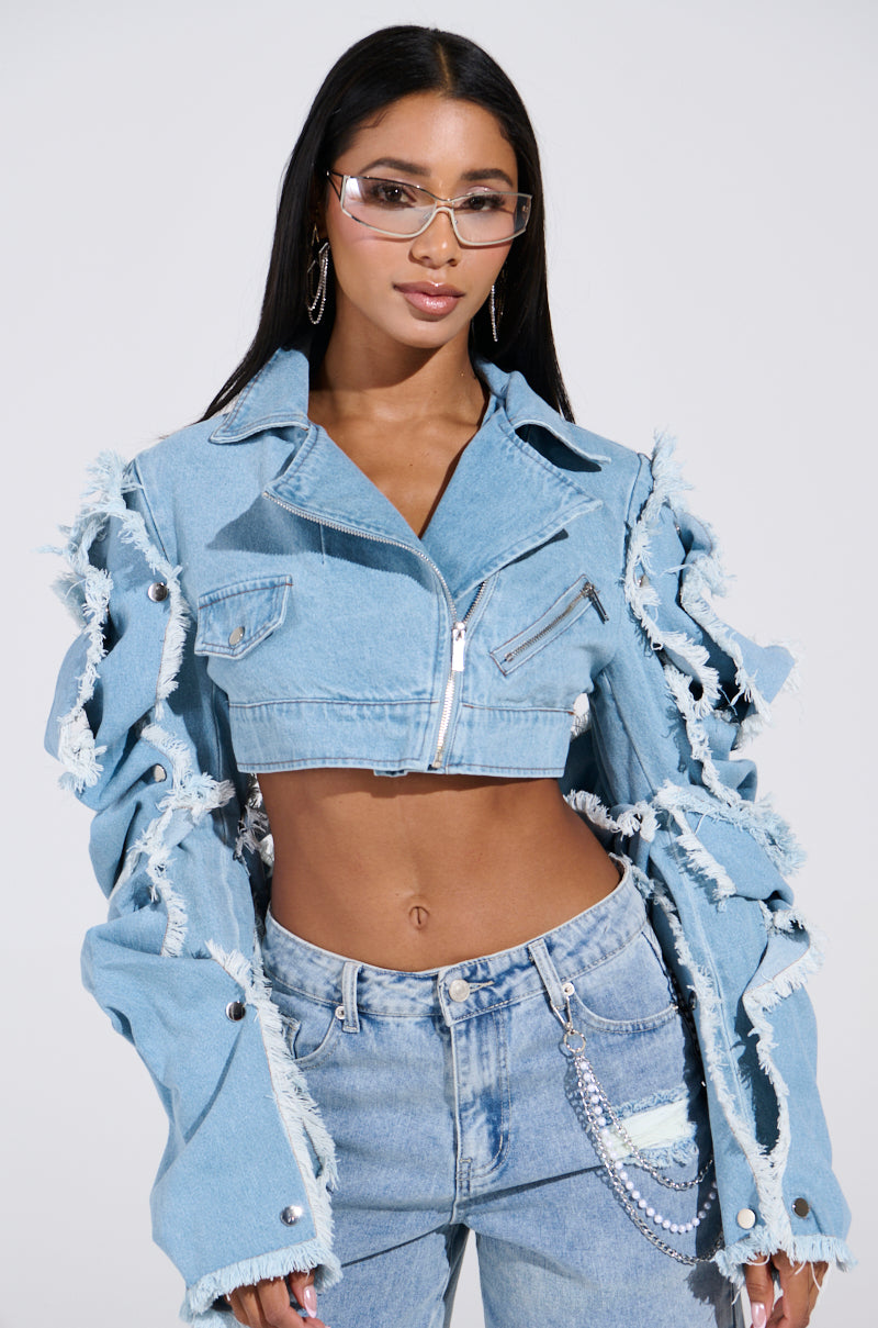 Wearing stylish glasses, an individual poses in the "SNAP OUT OF IT ULTRA CROP MOTO IN DENIM," a light wash jacket with frayed edges, zipper accents, and unique sleeve designs, paired with matching jeans. They stand confidently against a plain background.