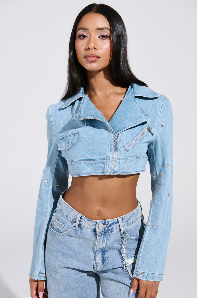 A person with long dark hair wears the SNAP OUT OF IT ULTRA CROP MOTO in light blue denim and pairs it with high-waisted jeans featuring a chain detail, standing against a plain gray background.