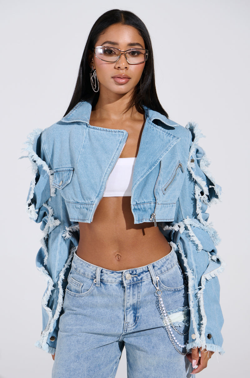 A person confidently wears the SNAP OUT OF IT ULTRA CROP MOTO IN DENIM over a white crop top and light wash jeans. They have long hair, wear glasses, rest one hand casually in their pocket, and sport a chain accessory dangling from the denim.