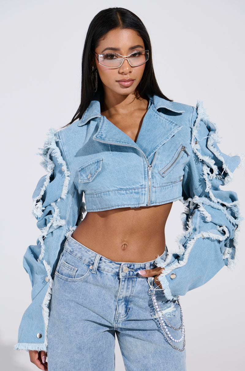 Someone sports the "SNAP OUT OF IT ULTRA CROP MOTO IN DENIM," a blue, ripped jacket with frayed edges and long sleeves. Paired with light wash jeans, transparent glasses, and chains, it exudes cool vibes in this minimalistic setting.