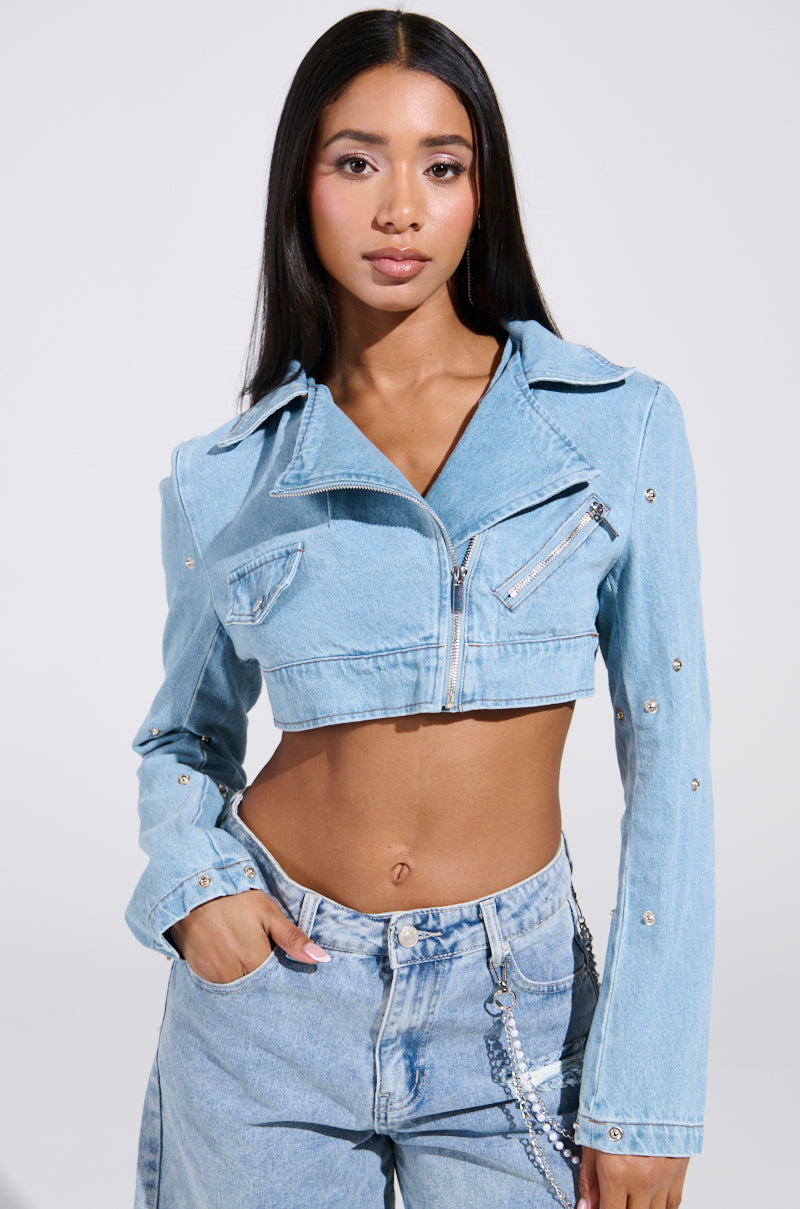 A person with long hair dons the SNAP OUT OF IT ULTRA CROP MOTO IN DENIM, featuring a light blue crop moto jacket and matching jeans. Their neutral expression contrasts against a plain light backdrop, highlighting their ultra-cropped, stylish look with one hand in a pocket.