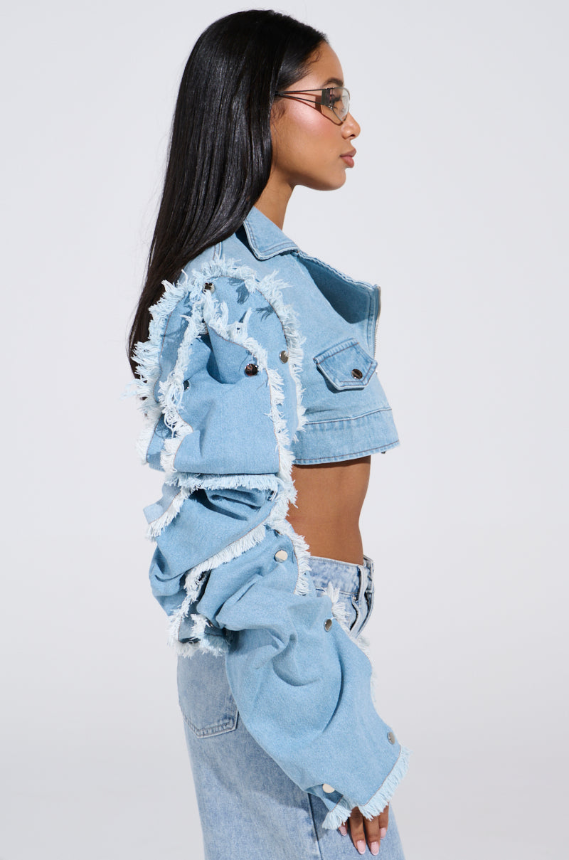 A long-haired person with glasses models the "Snap Out Of It Ultra Crop Moto in Denim," featuring a light wash, frayed edges, and button details. The ultra-cropped fit pairs perfectly with matching high-waisted jeans against a plain white backdrop.