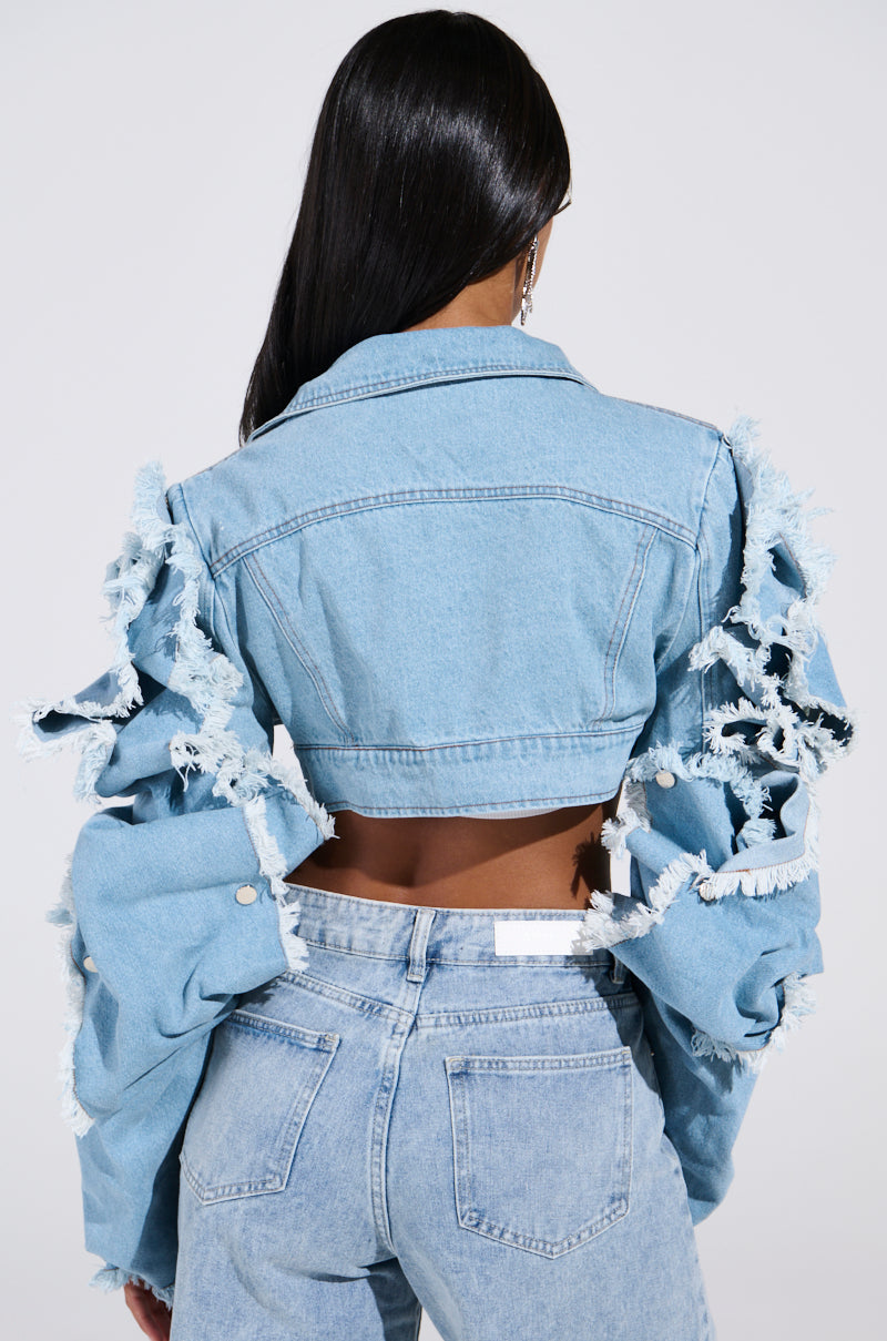 A rear view shows a person with long hair wearing the SNAP OUT OF IT ULTRA CROP MOTO IN DENIM, a light blue ultra-crop denim jacket featuring frayed edges and ruffled sleeves. They're paired with matching light wash jeans for a chic, effortless style.