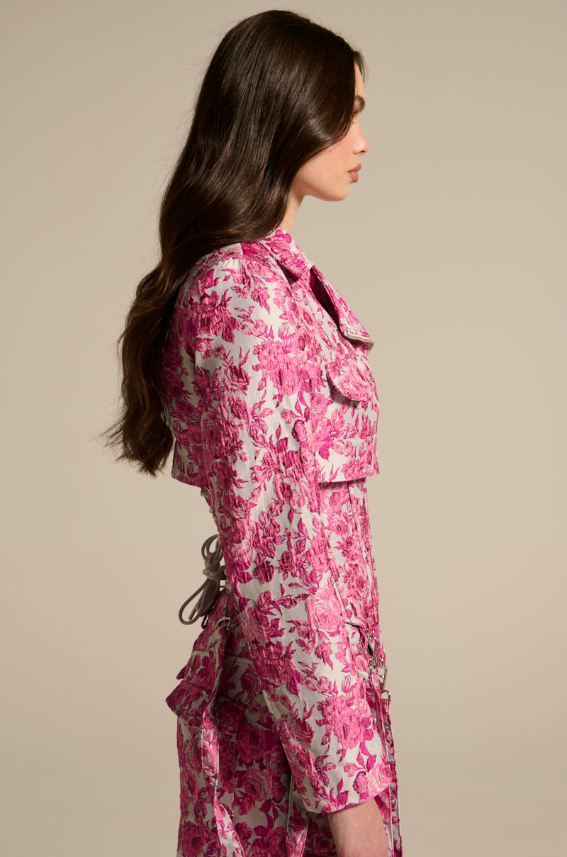 A woman with long brown hair in profile wears the Miley Jacquard Cropped Moto, a pink and white floral-patterned dress with a cinched waist. The outfit exudes sophistication with satin-lined touches against a neutral background.