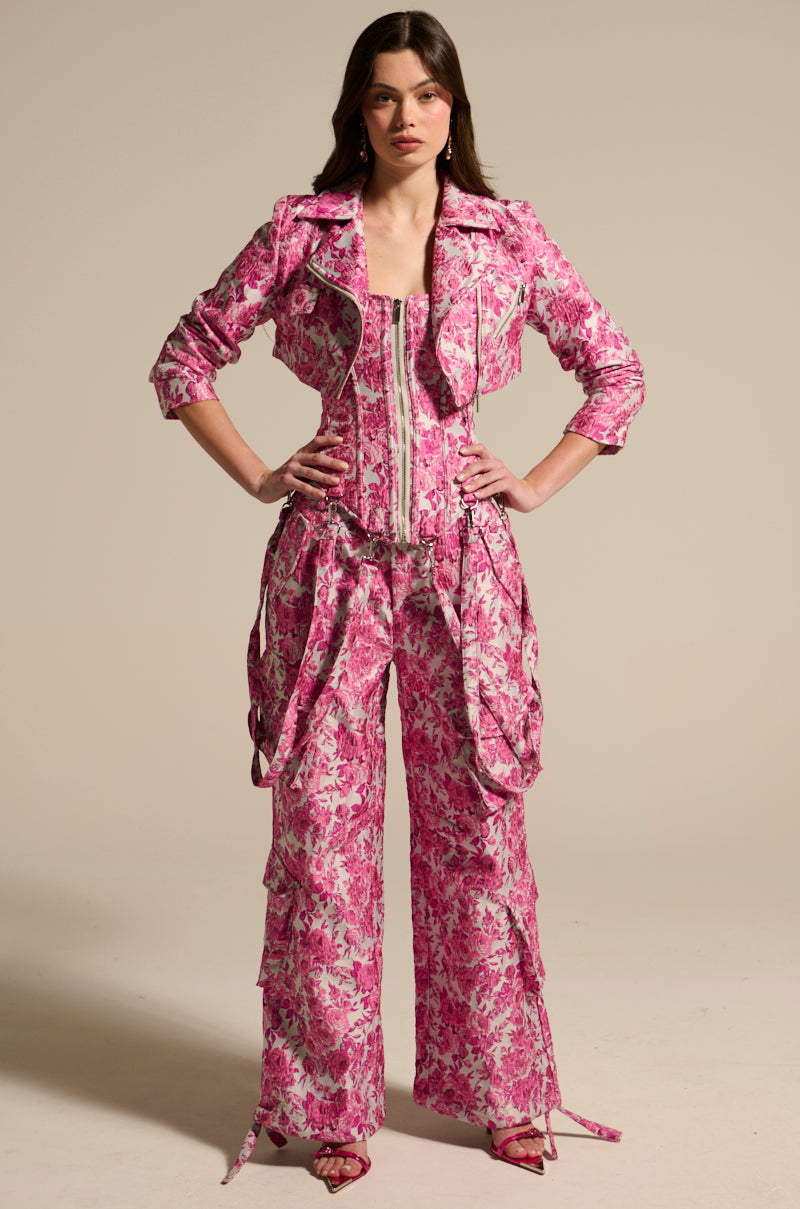 A person poses against a neutral background, wearing a vibrant pink floral outfit featuring the MILEY JACQUARD CROPPED MOTO jacket, corset, and wide-legged pants. They complement the look with matching pink high heels and wear their long hair loose.