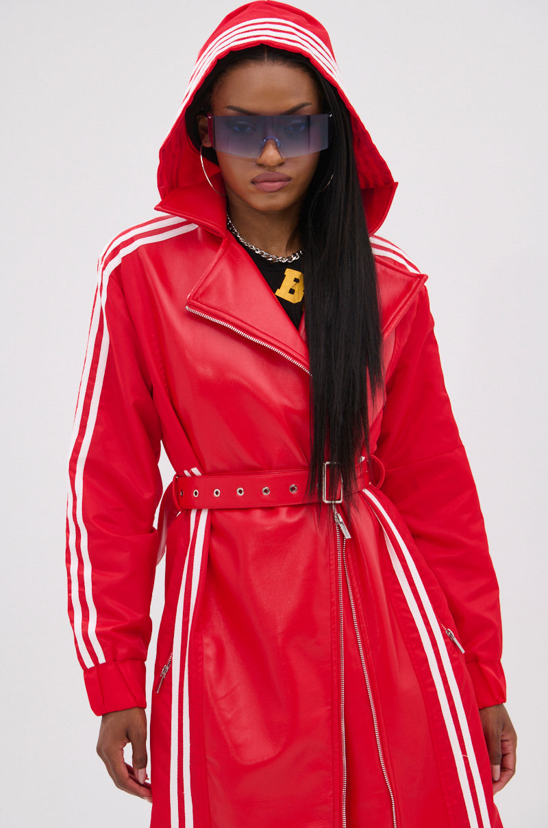 A person wears the UNAVAILABLE MIXED MEDIA TRENCH, a bright red faux leather coat with white racing stripes, paired with large blue-tinted sunglasses and a gold necklace, standing against a plain light background.