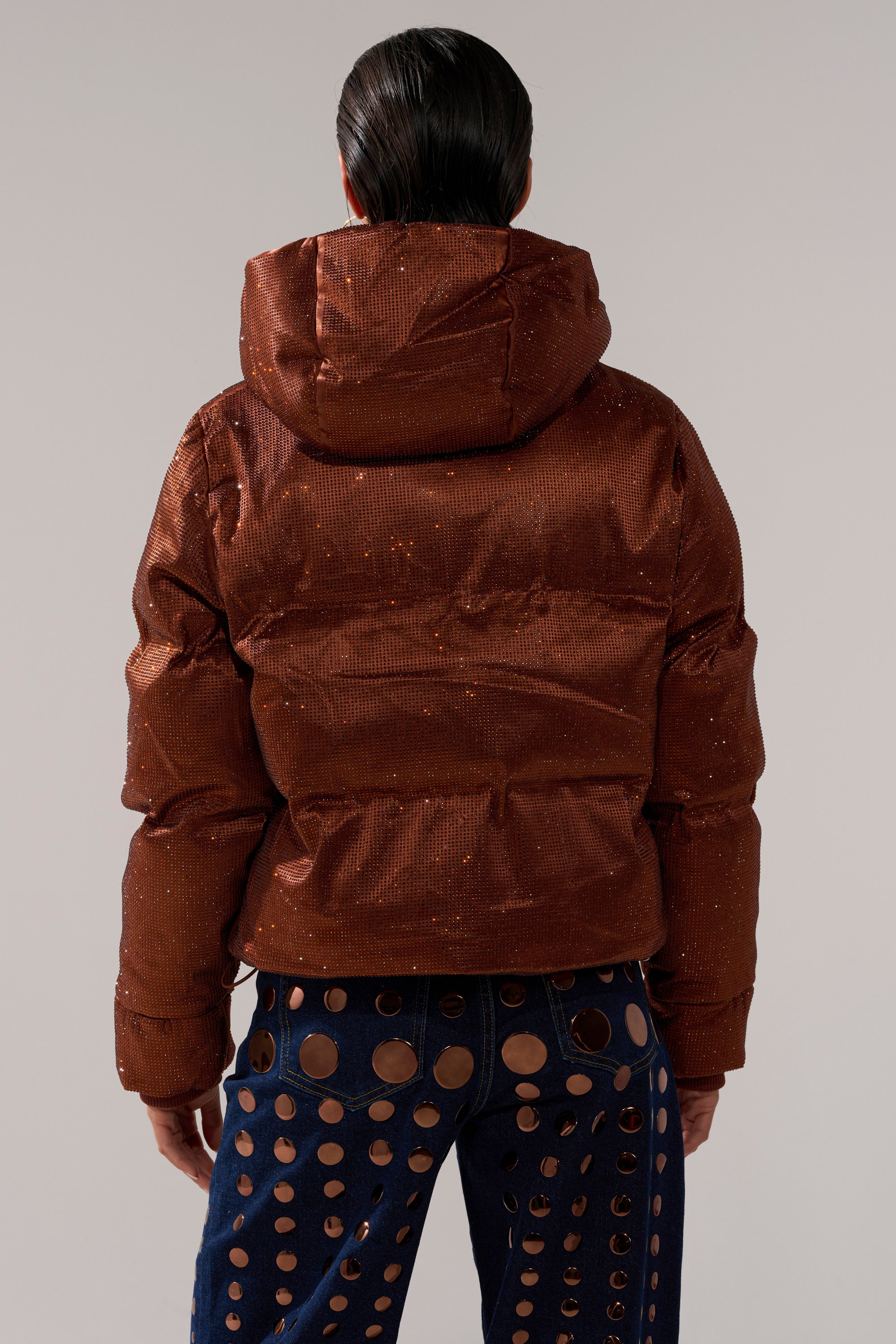 A person with slicked-back hair wears the FAYE RHINESTONE PUFFER IN COPPER and blue pants with large circular cutouts, viewed from behind against a plain background.